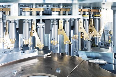Das Bild zeigt eine industrielle Blasformmaschine mit mehreren PET-Rohlingen, die in mechanischen Halterungen befestigt sind und auf den Blasvorgang vorbereitet werden.