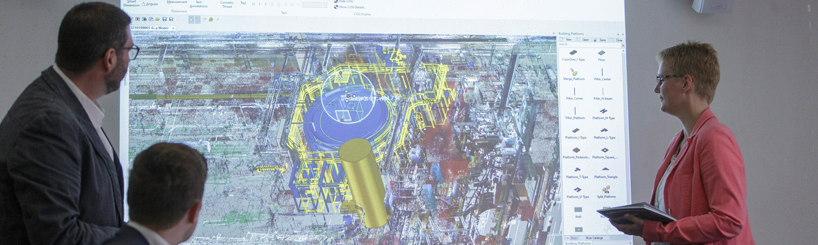 Drei Personen besprechen eine komplexe technische Zeichnung, die auf eine Leinwand projiziert ist, und analysieren dabei verschiedene Plattformtypen und Baupläne.