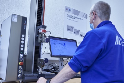 Ein Mann in einem blauen Pullover und einer Gesichtsmaske überwacht die Messdaten auf einem Computerbildschirm, während er ein Prüfgerät bedient.