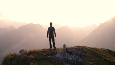 Eine Person steht auf einem Berggipfel und blickt in die Ferne, während die Sonne hinter den umliegenden Bergen aufgeht.